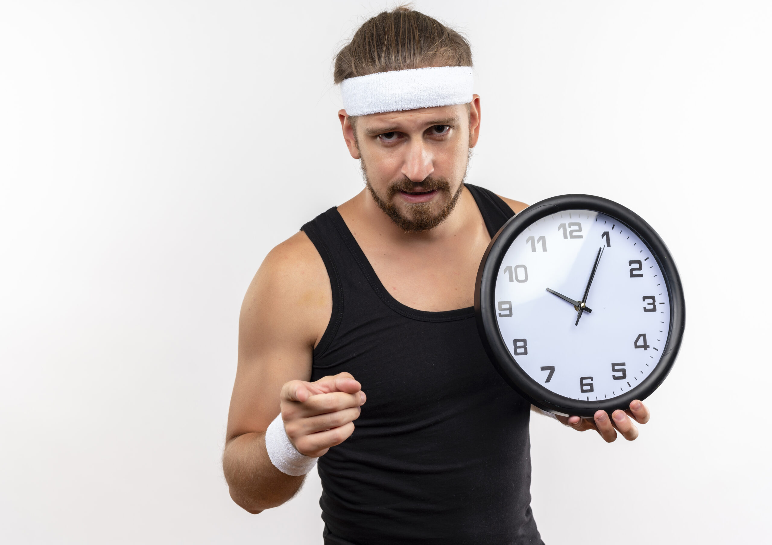 confident young handsome sporty man wearing headband and wristbands holding clock and pointing at camera isolated on white background with copy space